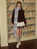 Full body shot of a woman in a white vest over a white long-sleeved shirt, a white pleated skirt, grey polka dot tights, silver boots, and a grey plaid scarf.