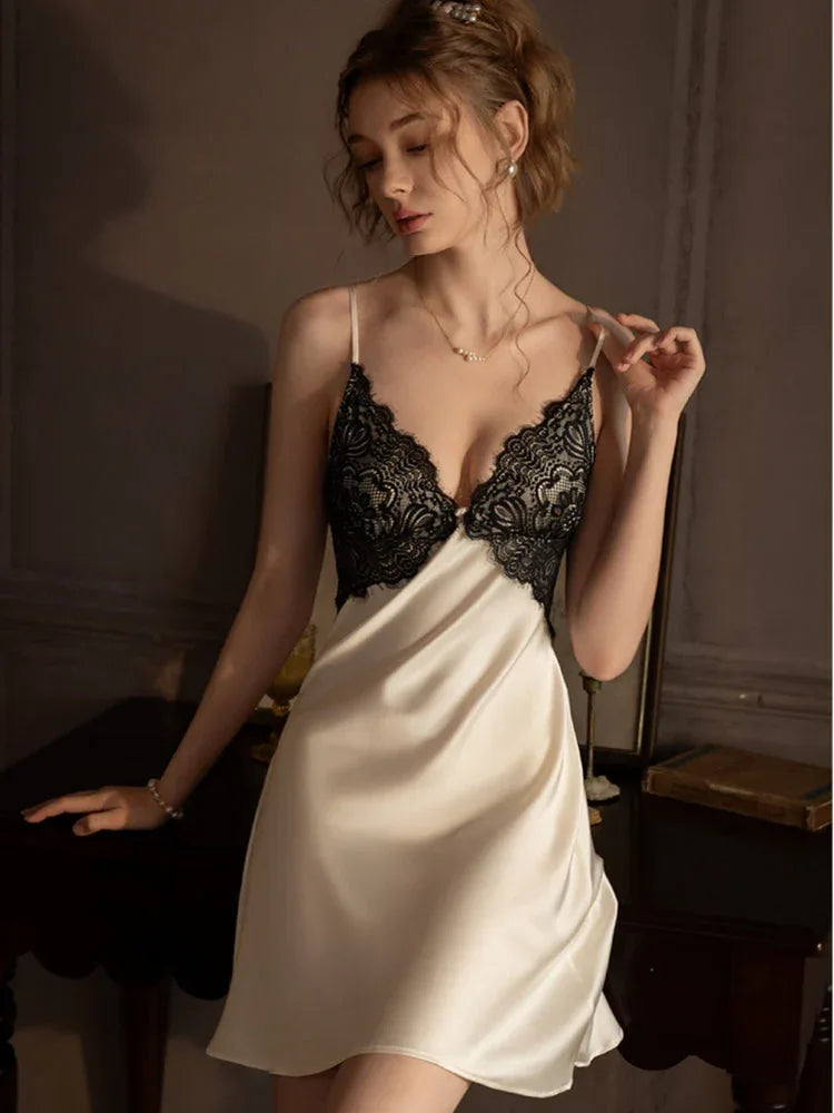 Woman in a cream satin chemise with black lace trim and spaghetti straps, posing with one hand on a table.