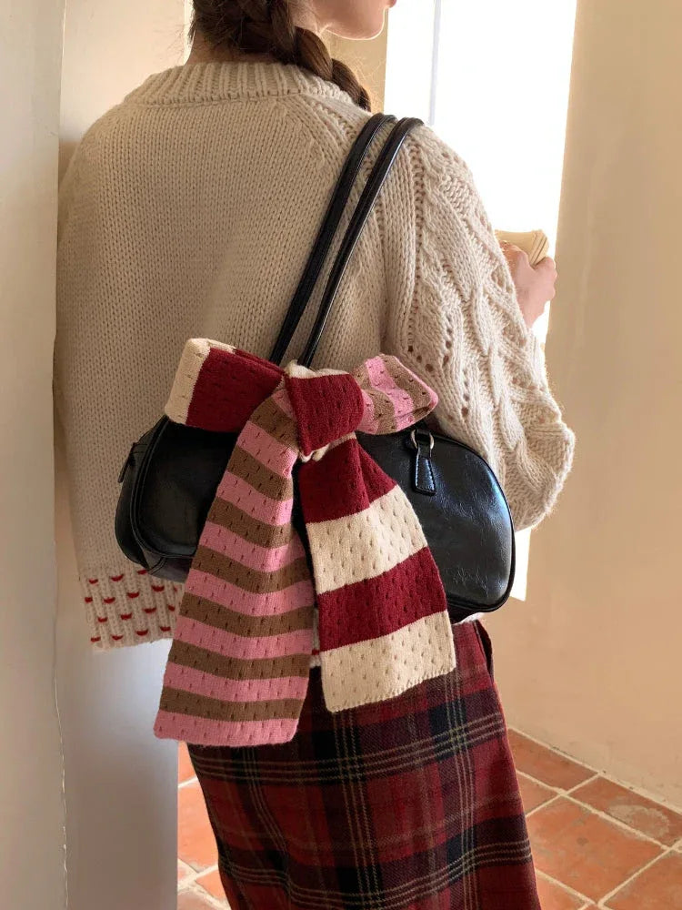 Rear view of a woman wearing a cream cable knit cardigan and plaid skirt, with a striped scarf tied to a black handbag.