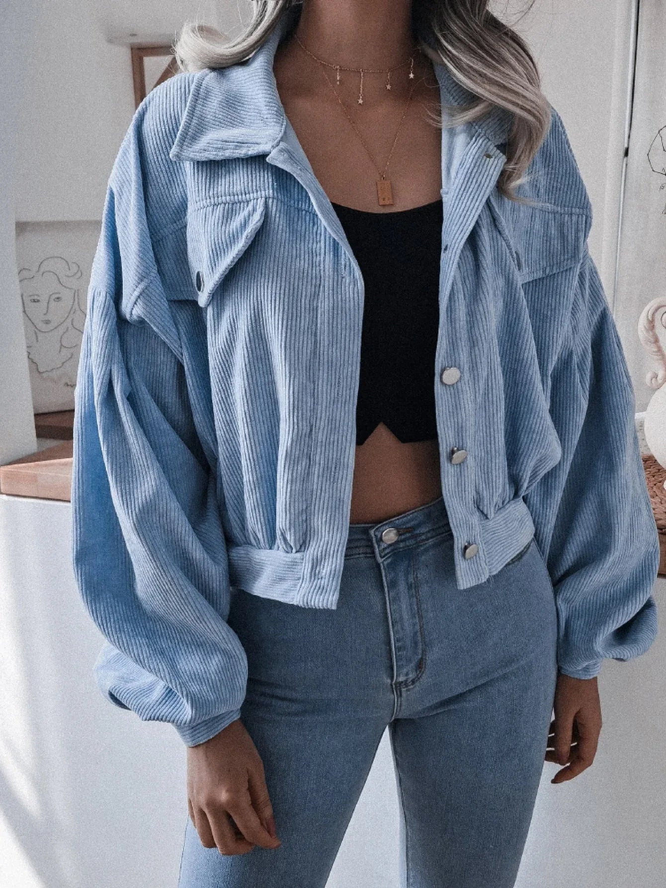 Woman wearing a light blue corduroy cropped jacket with balloon sleeves and a collared neckline, paired with a black crop top and blue jeans.