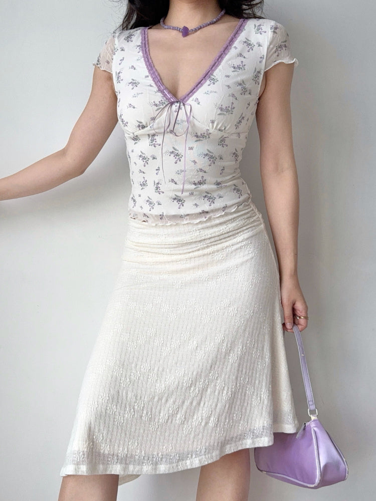 Full body shot of a woman wearing a white floral print mesh top and a cream-colored textured skirt. She is accessorized with a purple beaded necklace with a butterfly pendant and a small purple handbag.