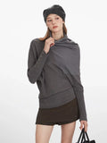 Close-up front view of a woman in a grey asymmetrical knit sweater with draped overlay, adjusting the neckline.