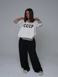 Woman wearing a white knit sweater with CCCP graphic print and black stripes on cuffs and hem.