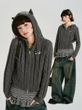 Model wearing a grey cable knit cropped hooded jacket with ear details, paired with striped top and jeans.