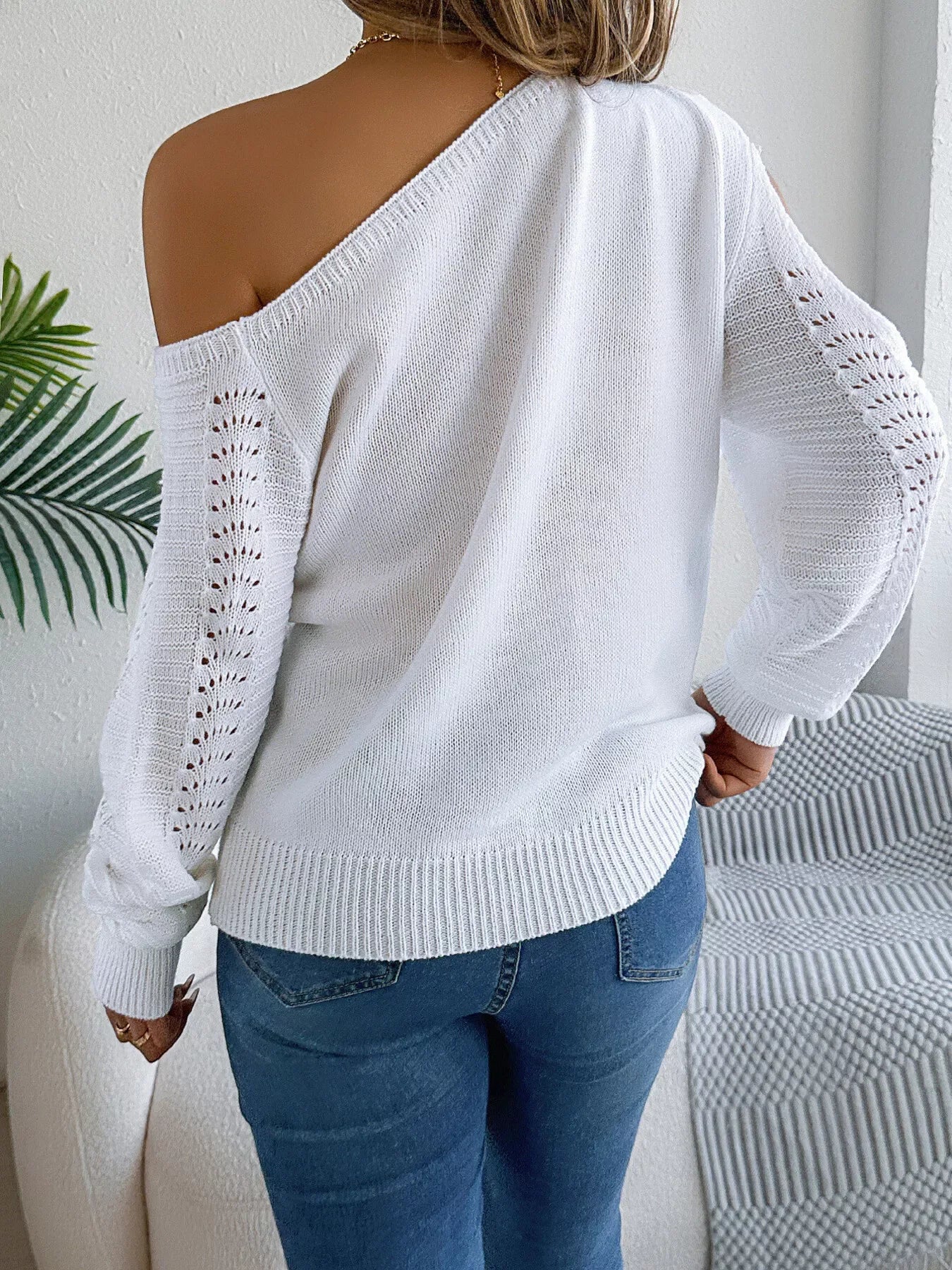 Back view of a woman in a white off-the-shoulder knit sweater with cut-out sleeves and blue jeans