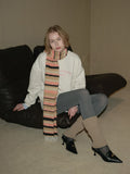 Woman wearing a cream-colored sweatshirt, grey leggings, leg warmers, a multi-colored striped scarf, and black loafers sitting on a black couch.
