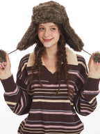 Close-up of a woman wearing a brown striped knit sweater with a v-neck and collar detail, and a brown faux fur hat with ear flaps, adjusting the hat's ear flaps.
