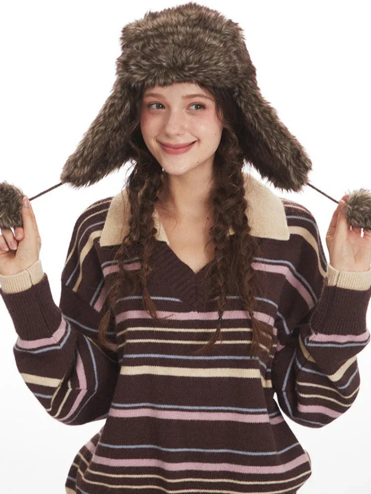 Close-up of a woman wearing a brown striped knit sweater with a v-neck and collar detail, and a brown faux fur hat with ear flaps, adjusting the hat's ear flaps.