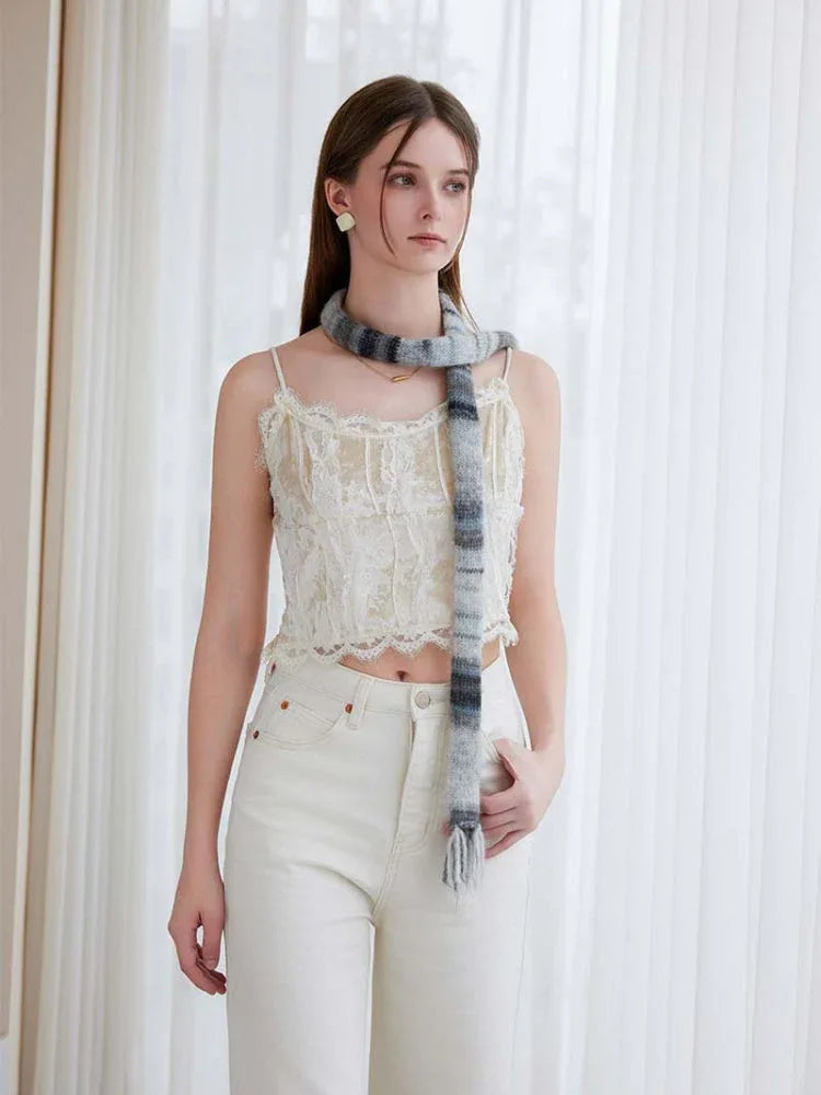 Model posing with hands in pockets, wearing a white lace camisole and cream trousers, with a striped knit scarf.