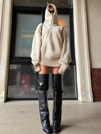 Woman posing outdoors in a beige hooded knit sweater and black leather boots.