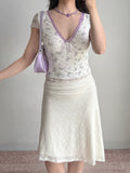 Woman posing with her hand on her hip, wearing a white floral print mesh top and a textured cream skirt. She holds a purple handbag and wears a purple beaded necklace.