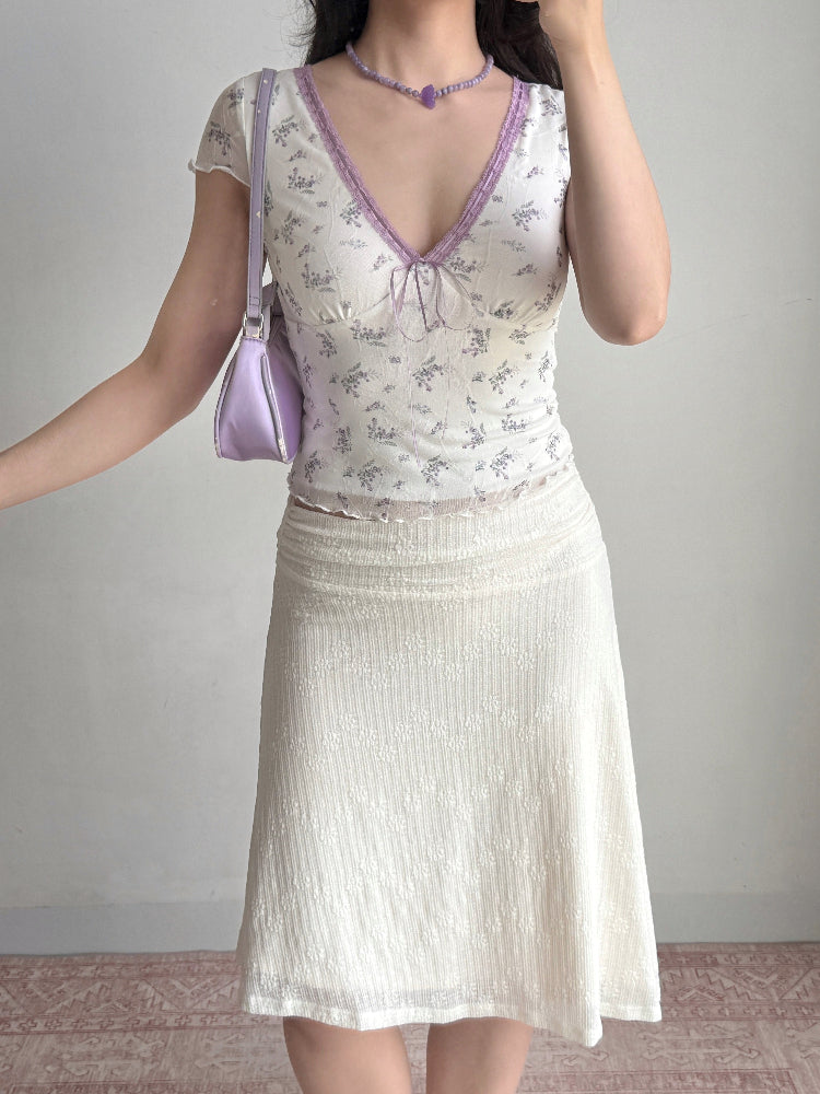 Woman posing with her hand on her hip, wearing a white floral print mesh top and a textured cream skirt. She holds a purple handbag and wears a purple beaded necklace.