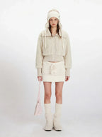 Woman wearing a cream fuzzy cropped jacket and matching mini skirt, with a knitted hat and boots
