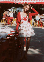 Full body shot of a woman in a white tiered mini dress with red trim and bow detail, standing outdoors