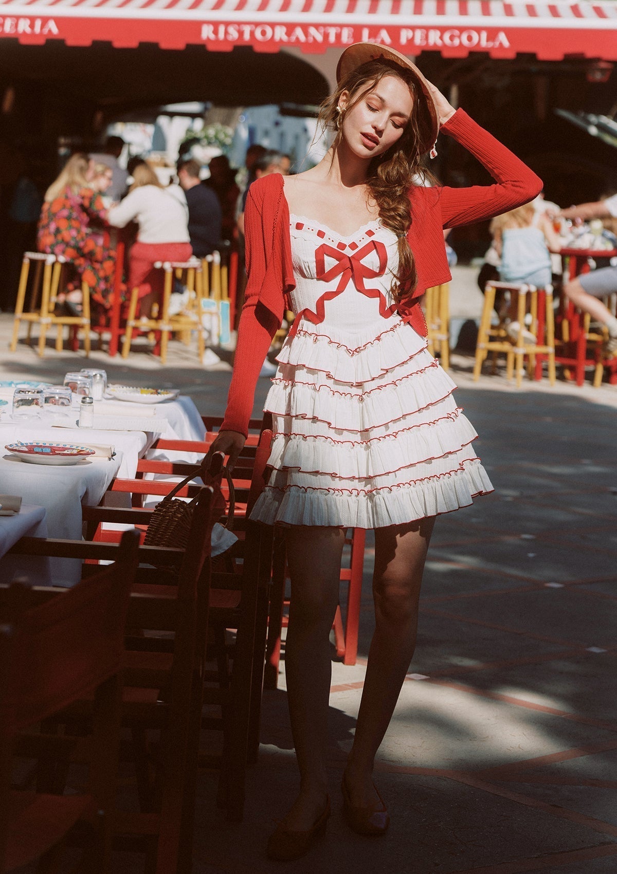 Full body shot of a woman in a white tiered mini dress with red trim and bow detail, standing outdoors