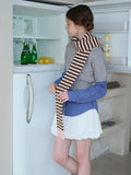 Woman wearing a grey cropped sweater over a blue gingham shirt, white bubble shorts, navy polka dot socks, white Mary Jane shoes, and a pink and brown striped scarf, looking inside a refrigerator.