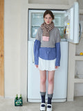 Full body shot of a woman wearing a grey cropped sweater over a blue gingham shirt, white bubble shorts, navy polka dot socks, white Mary Jane shoes, and a pink and brown striped scarf.