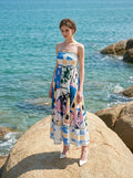 Woman wearing a white midi dress with an abstract botanical print and spaghetti straps, standing on a rock by the turquoise sea.