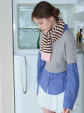 Woman wearing a grey cropped sweater over a blue gingham shirt, white bubble shorts, navy polka dot socks, white Mary Jane shoes, and a pink and brown striped scarf, looking down.