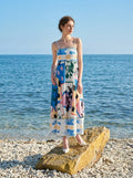 Woman wearing a white midi dress with an abstract botanical print and spaghetti straps, standing on a rock by the sea.