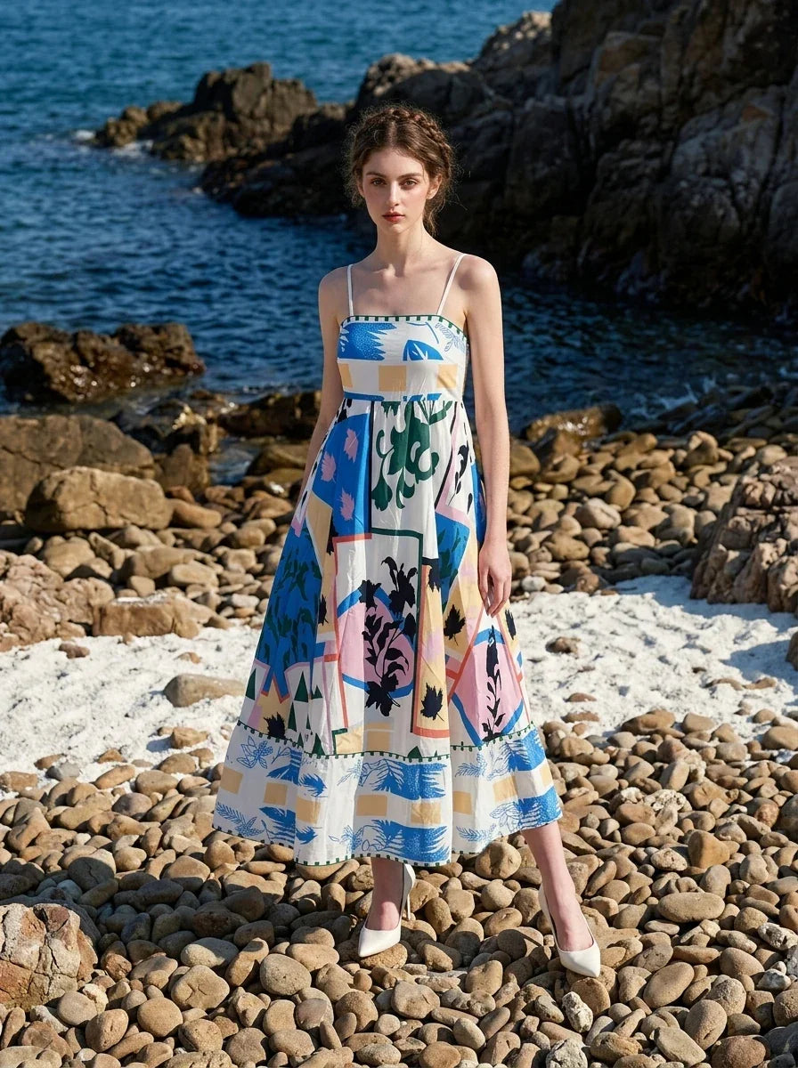 Woman wearing a white midi dress with an abstract botanical print and spaghetti straps, posing on a rocky beach.