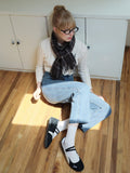 Woman sitting on a wooden floor wearing a cream cable knit cropped sweater, plaid scarf, blue jeans, and black and white flats.