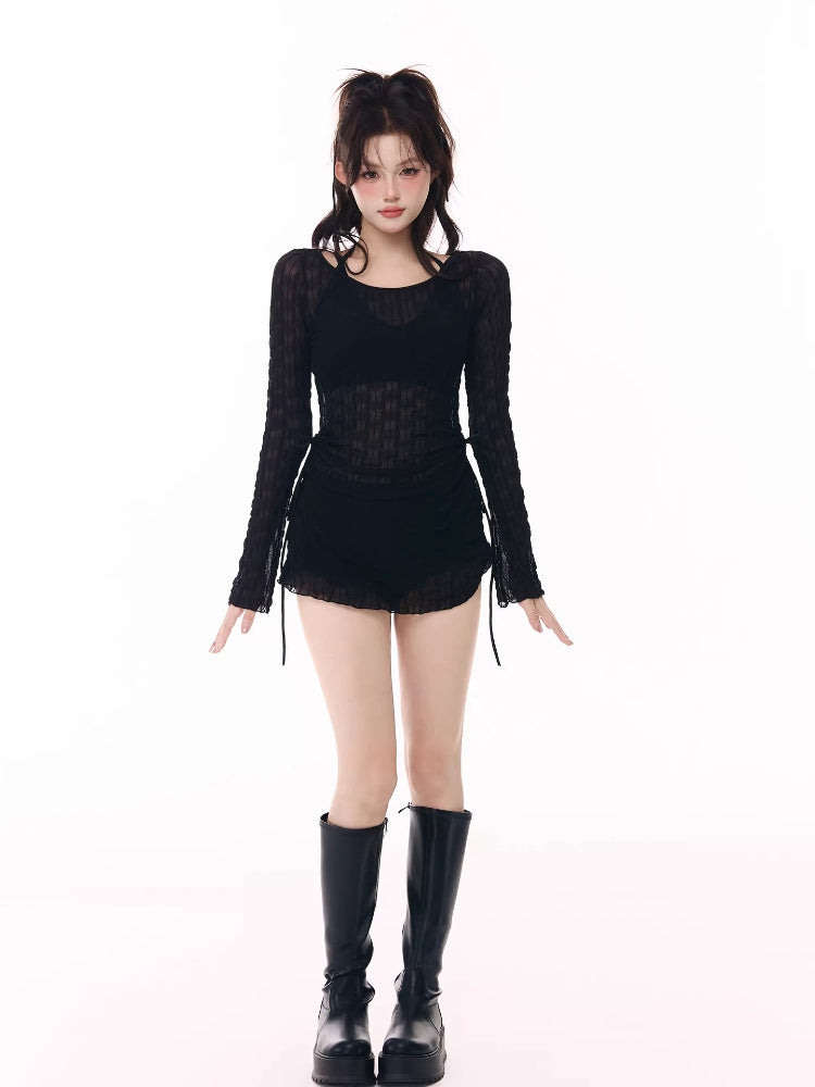 Woman wearing a black sheer long-sleeve top over a black bikini top and black ruffled hem shorts, with black platform boots.