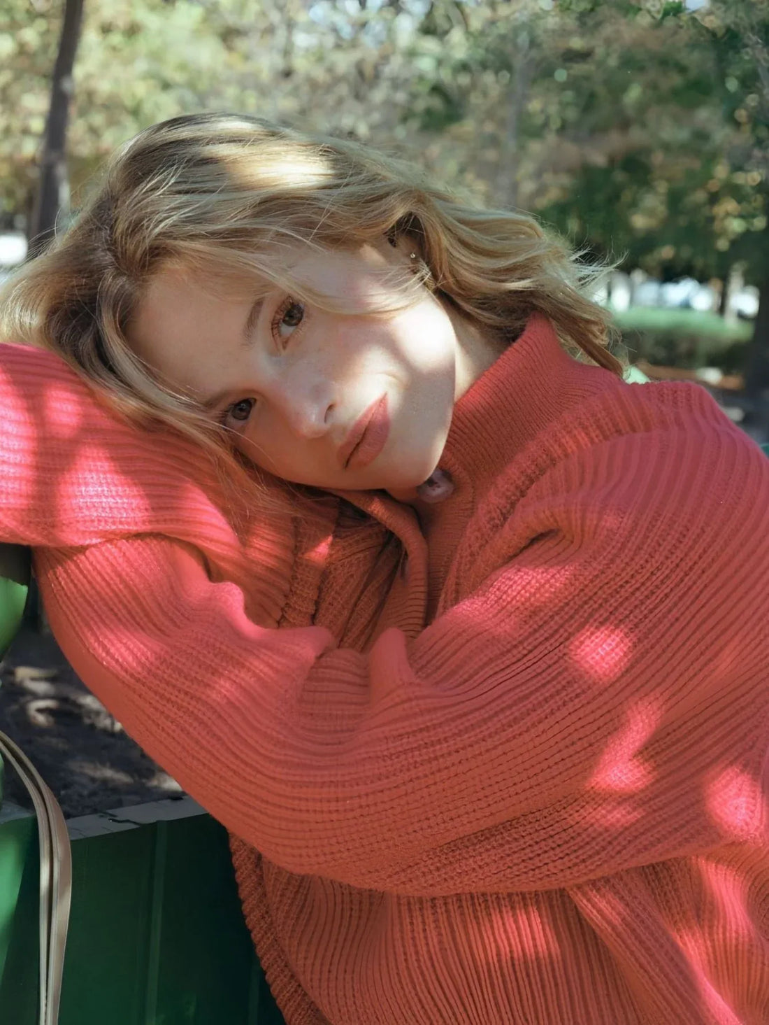 Woman wearing a coral knit sweater with a high neckline and button placket, paired with blue denim jeans, outdoors in a park.