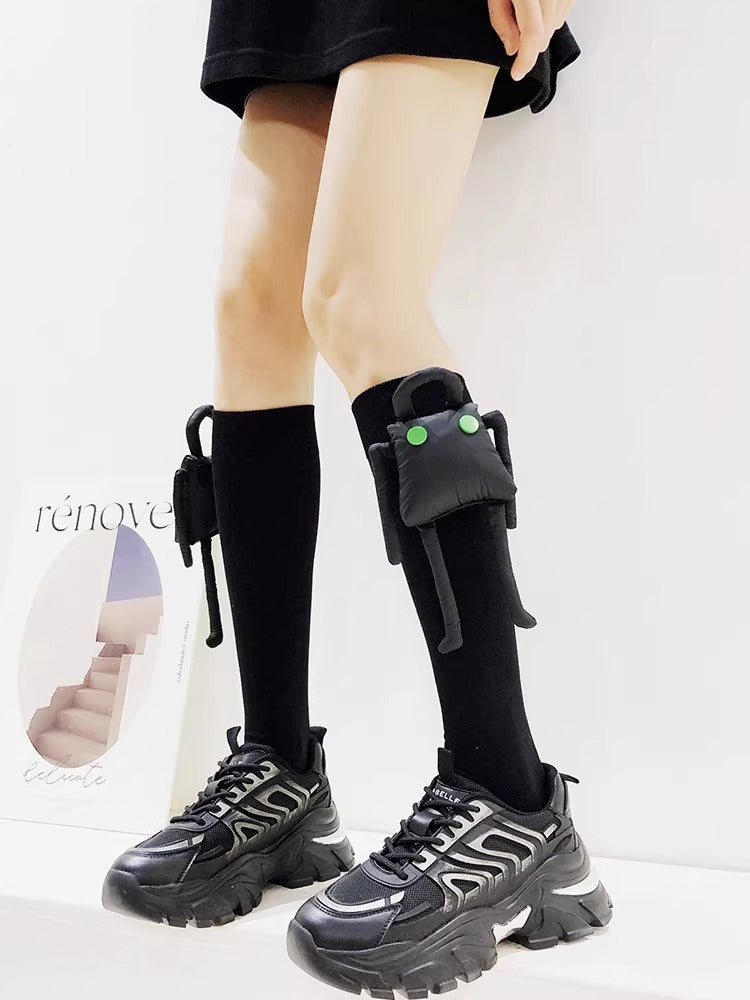 Black knee-high socks with a detachable plush toy embellishment on each leg, worn with black chunky sole sneakers.