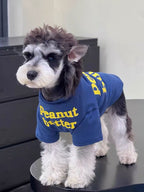 A small dog stands on a surface, wearing a blue t-shirt with yellow text.