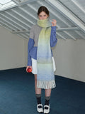 Woman wearing a long, oversized scarf with an ombre gradient of yellow, blue, and green, draped around her neck over a gray sweater and blue plaid shirt.