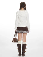 Back view of a woman wearing a white hooded zip-up jacket with a brown patterned skirt, white knee-high socks, and brown boots.
