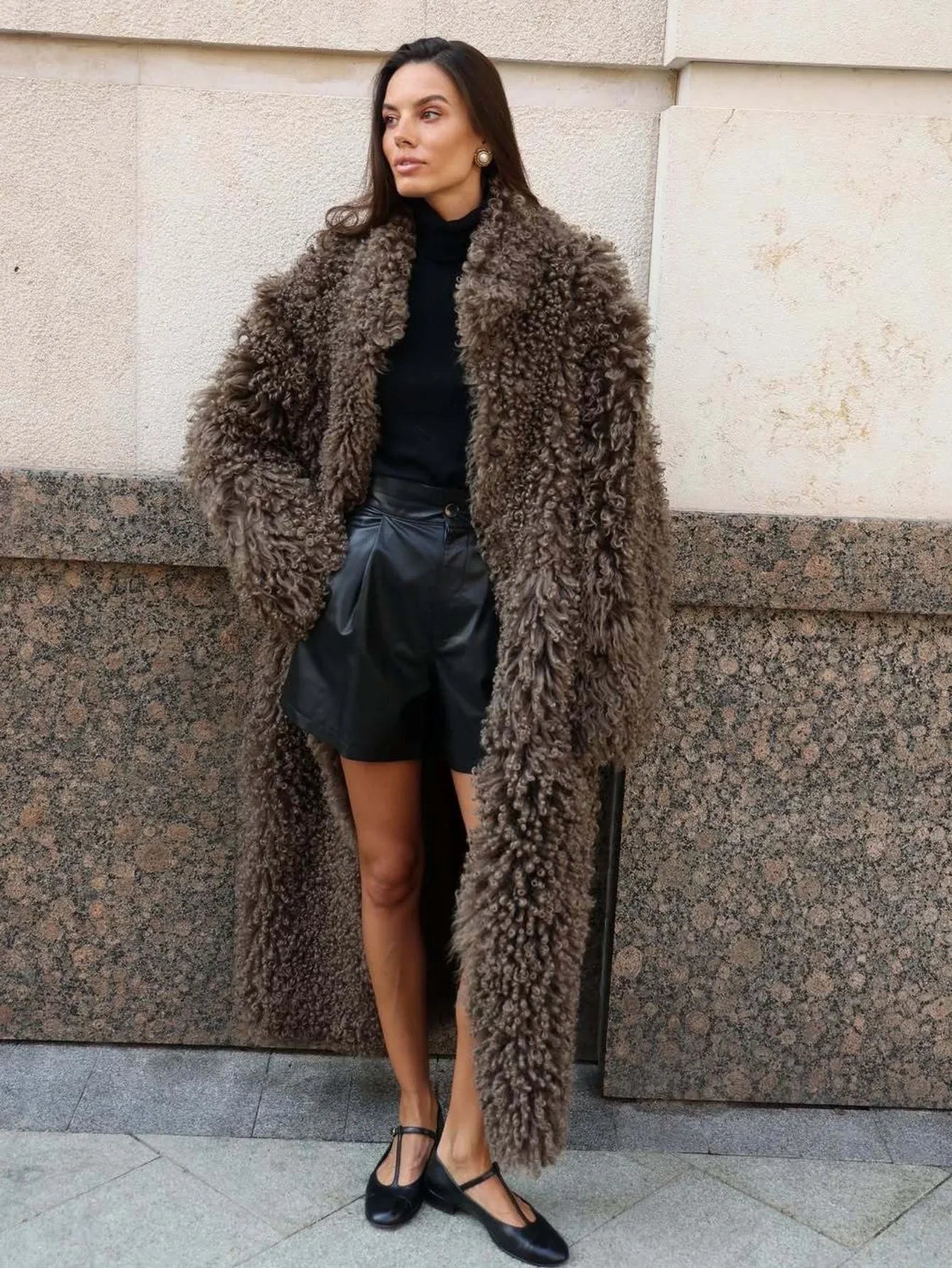 Woman wearing a long brown textured faux fur coat with a black turtleneck and leather shorts. Main image