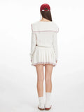 Back view of a woman in a white knit sweater with a navy sailor collar and red trim, and a white pleated skirt.