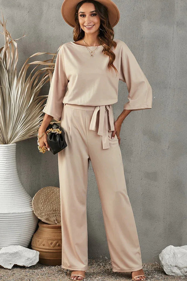 Woman wearing a beige wide leg jumpsuit with a boat neck and tie waist. Main image