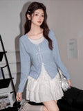 Full body shot of a woman in a light blue knit cardigan and white tiered skirt, holding a silver handbag.