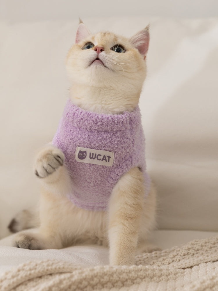 A light purple fluffy knit pet vest with a mock neck and a white logo patch on the front, worn by a light-colored cat.