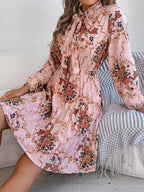 Woman sitting on a couch wearing a pink floral print midi dress with a bow neckline and pleated skirt
