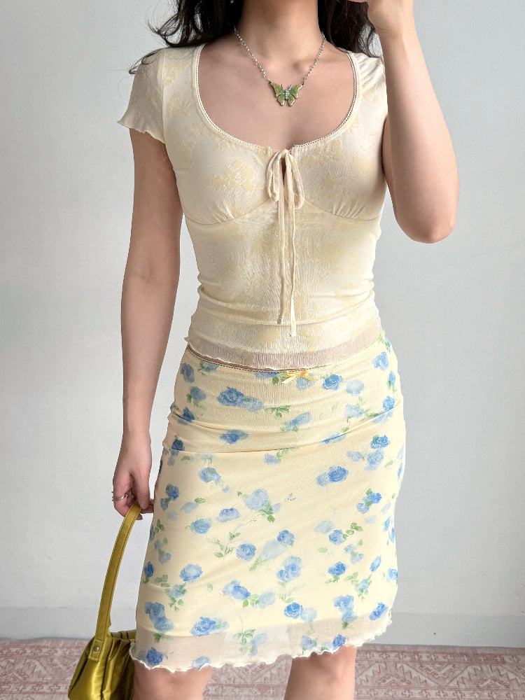 Full body shot of a woman in a yellow sheer floral top and a matching floral print skirt, holding a green handbag.