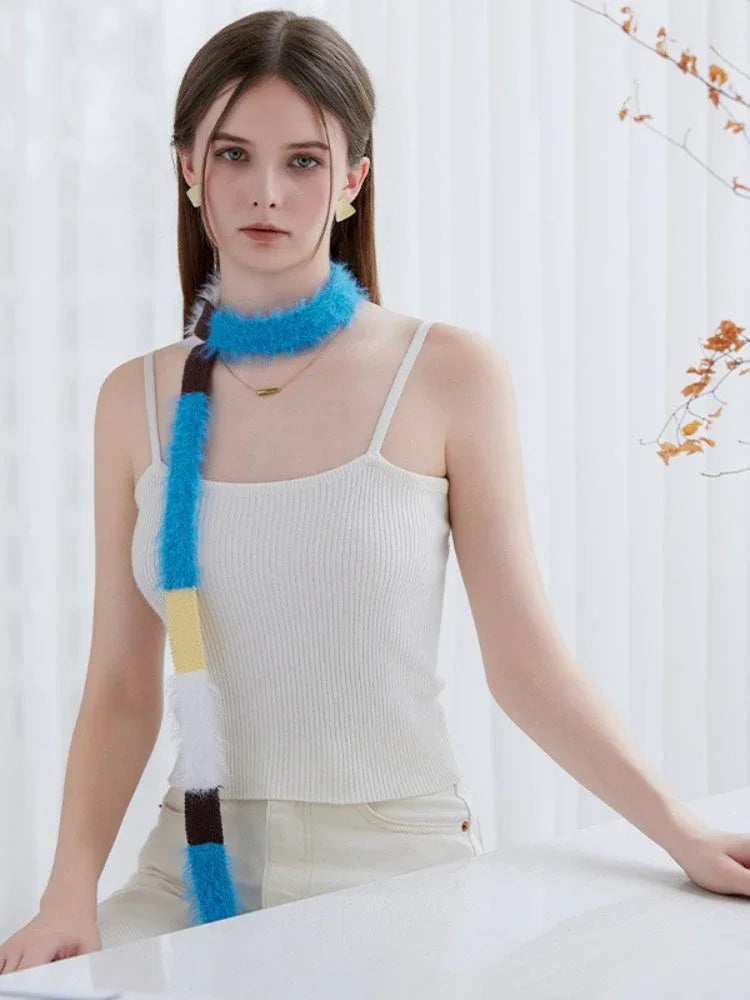 Woman wearing a white ribbed tank top and cream pants, accessorized with a long, fuzzy knit scarf featuring alternating blocks of blue, yellow, white, and brown.