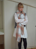 Model with arms crossed, wearing a plaid scarf with fringe trim over a white shirt and grey vest.