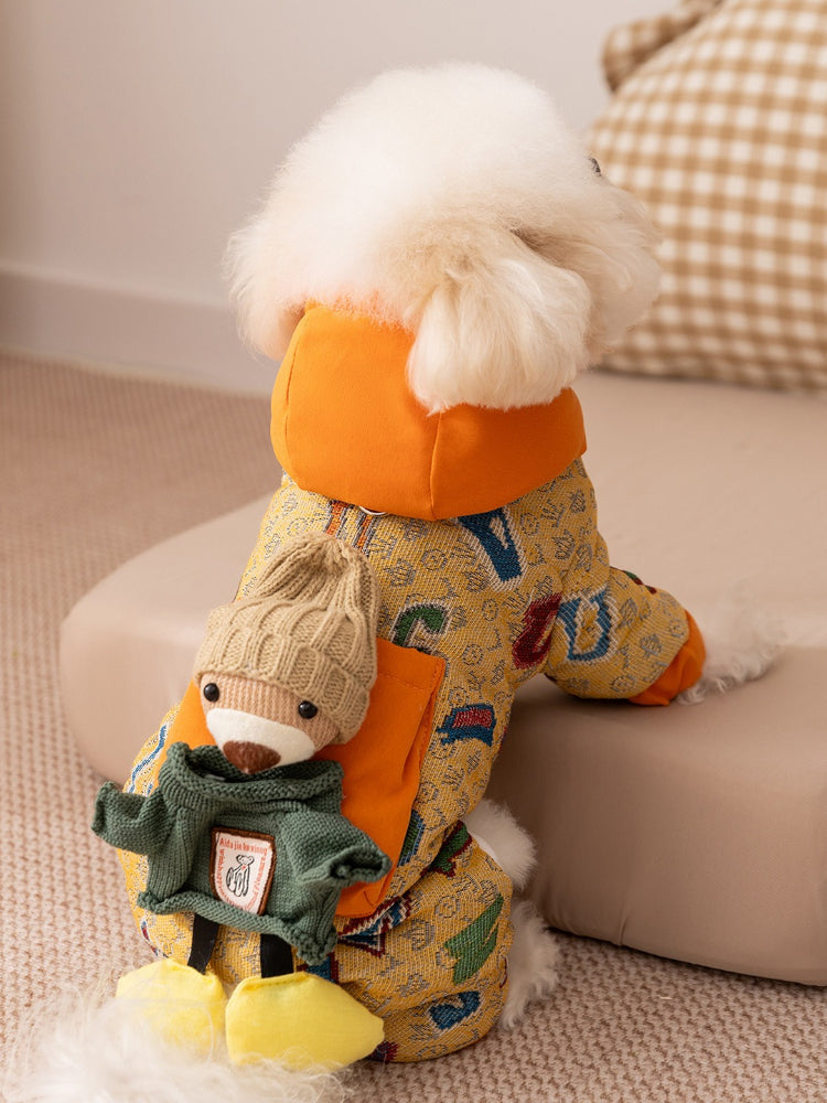 Rear view of a small white dog in a yellow patterned dog jacket with a teddy bear applique on the back.