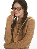 Close-up of a woman's face and upper body, wearing a brown hooded cable knit sweater