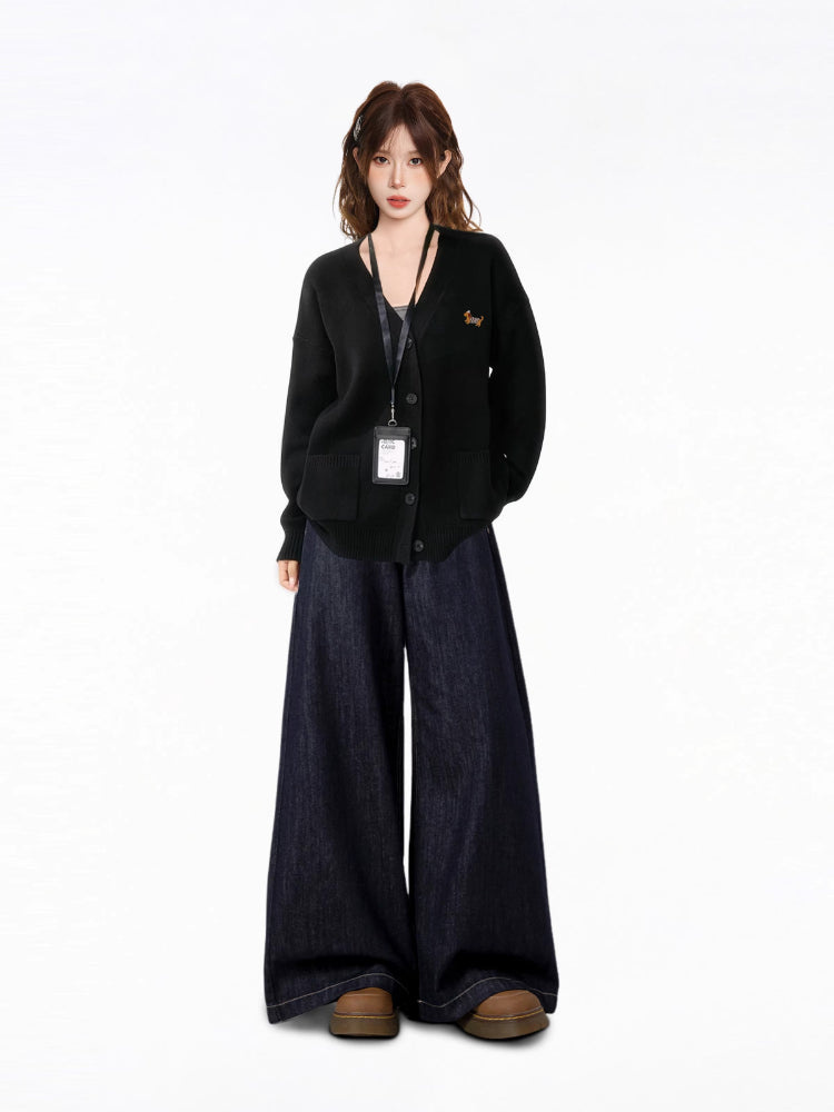 Woman standing in a relaxed pose, wearing a black oversized knit cardigan over a dark tank top, paired with wide-leg dark wash jeans and chunky brown shoes. An ID badge holder is visible.