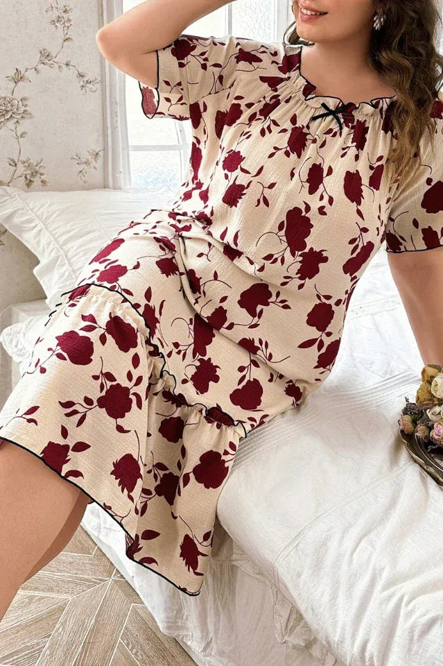 Plus-size woman wearing a cream-colored short-sleeve nightgown with a red floral print and ruffled hem, sitting on a bed.