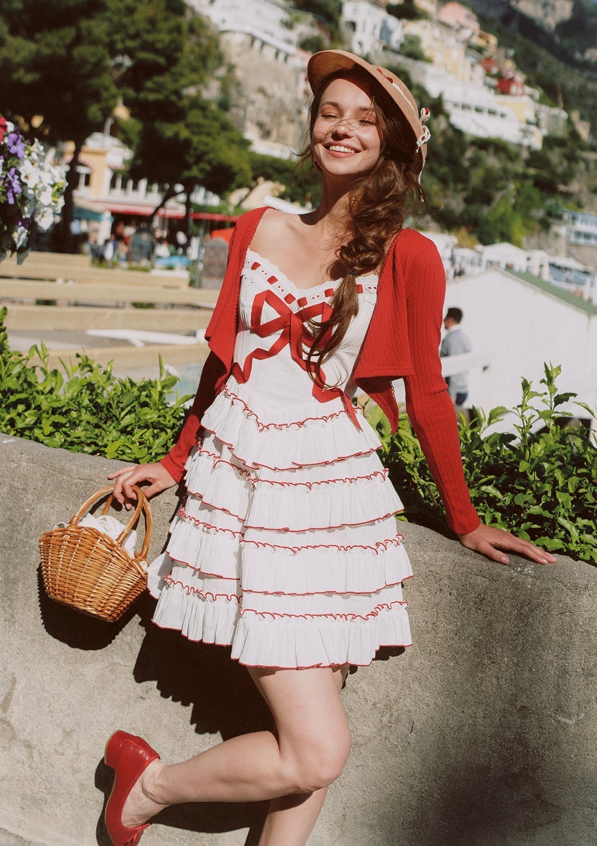 Model with braided hair wearing a white tiered mini dress with red trim and bow detail, a straw hat, and red shoes