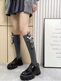 Woman standing, showcasing grey ribbed knee-high socks adorned with a plush bunny charm, worn with a pleated skirt and black platform shoes.