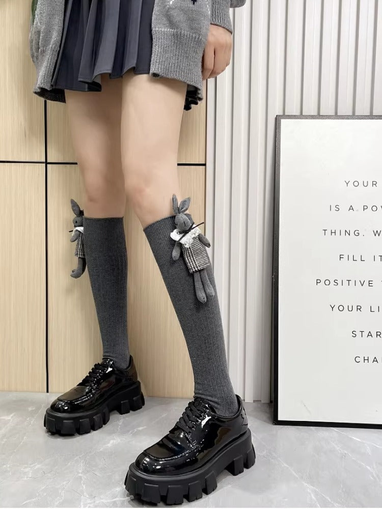Woman standing, showcasing grey ribbed knee-high socks adorned with a plush bunny charm, worn with a pleated skirt and black platform shoes.