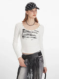 Woman wearing a white long-sleeve graphic t-shirt with a scoop neckline and studded details, posing with hands on head.