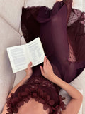 Woman lying on a sofa reading a book, wearing a plum slip dress with sheer overlay and floral trim.